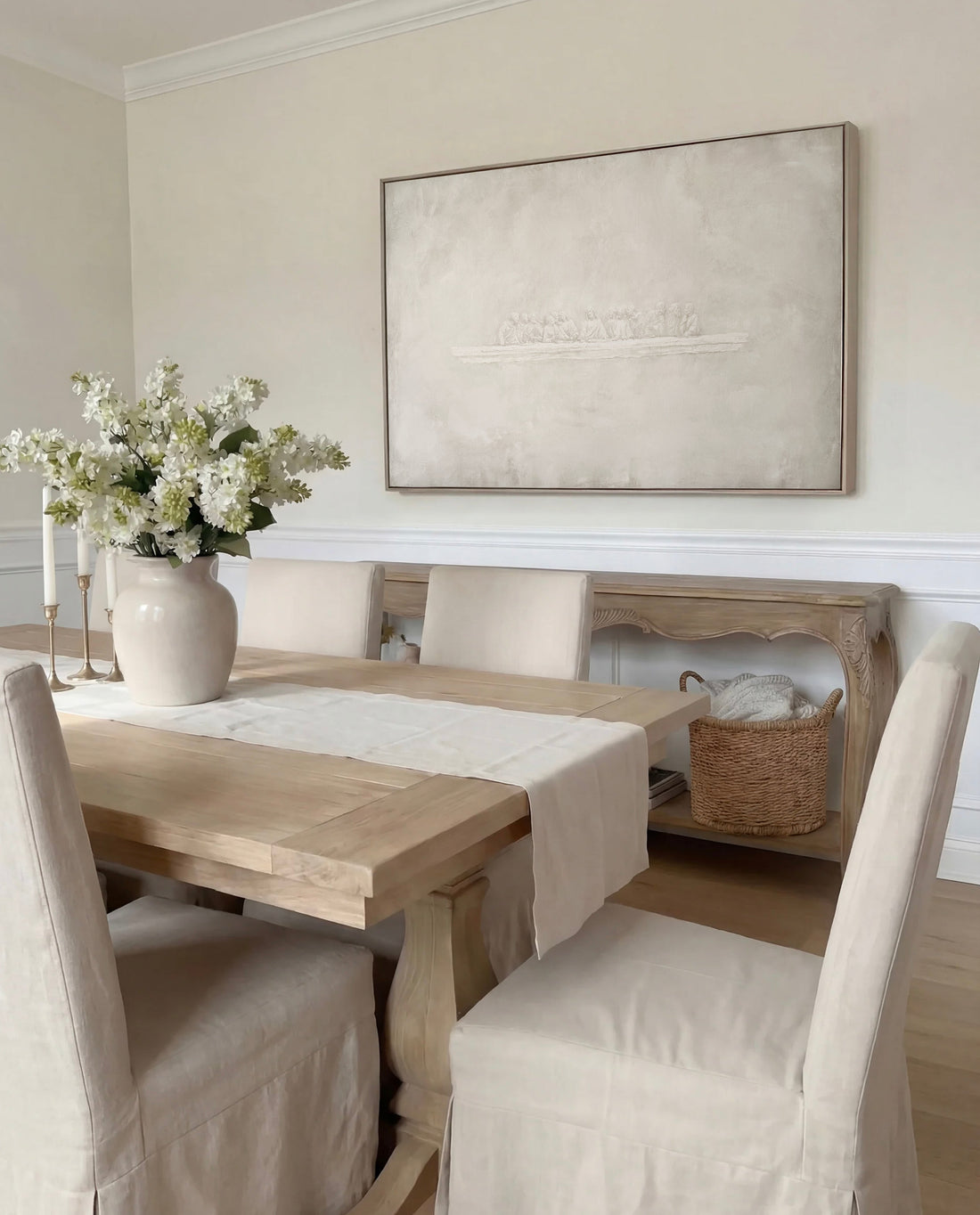 The Last Supper canvas in a light wood frame hung above a dining table with slipcovered chairs and a vase of white flowers